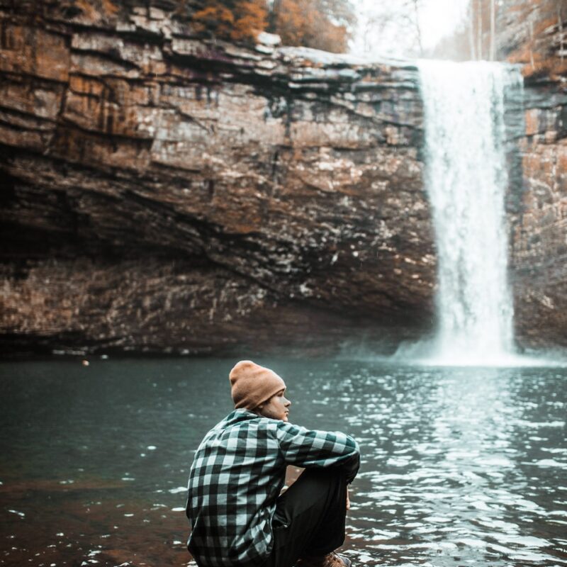 man sitting on brown stone near waterfall during daytime